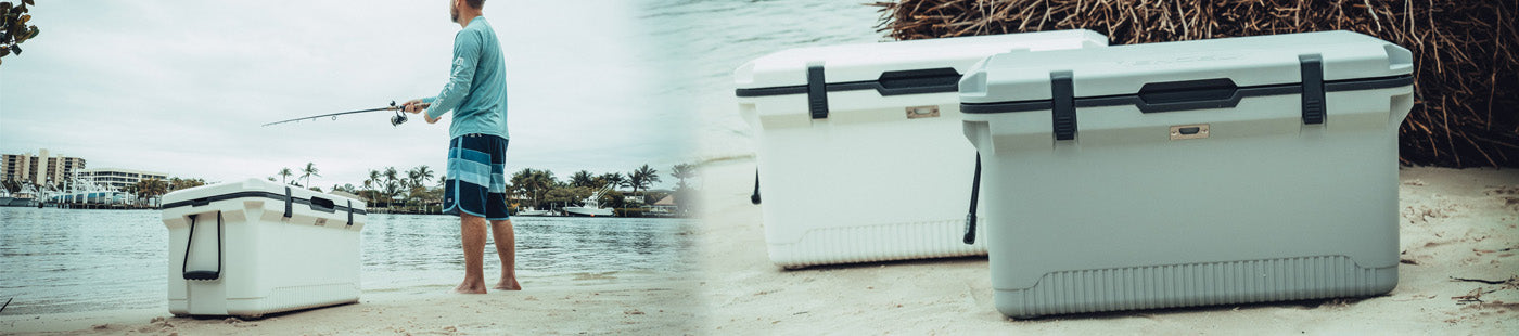 A person fishing by the water stands near two large white coolers placed on sandy ground with plants and buildings in the background.