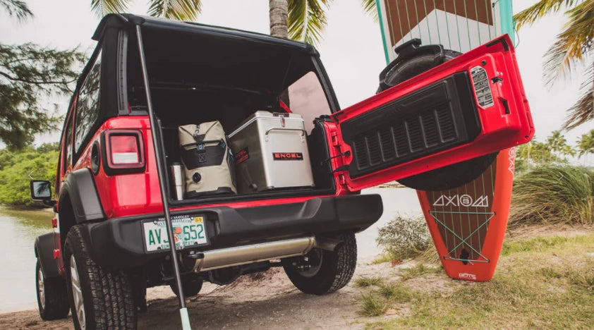 Red suv with open trunk displaying a cooler and a surfboard near a palm tree.