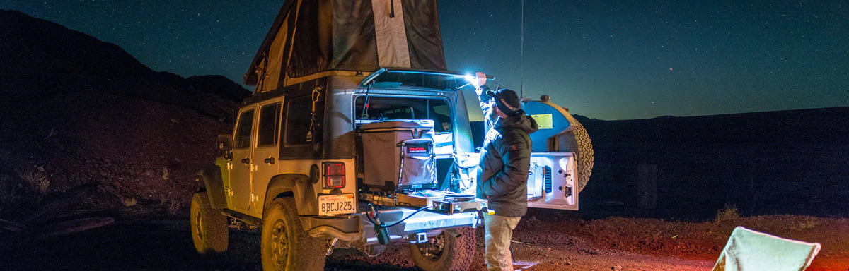 Person stands at the back of a Jeep with a rooftop tent, preparing food or gear at night under a hanging light in an outdoor, remote setting.