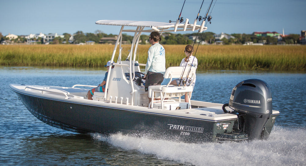 Two people operate a black Pathfinder 2200 boat with fishing rods, traveling on the water near a grassy shoreline and houses in the background.