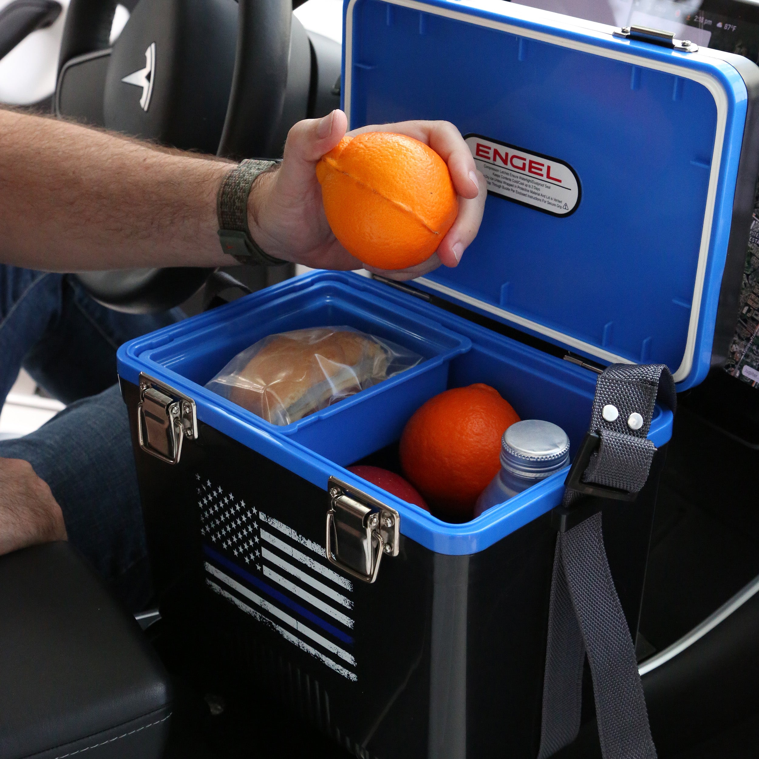 In a car, someone holds an orange near the Engel Coolers 7.5Qt Thin Blue Line Lunch Box. Inside, there's a sandwich, two more oranges, and a drink.