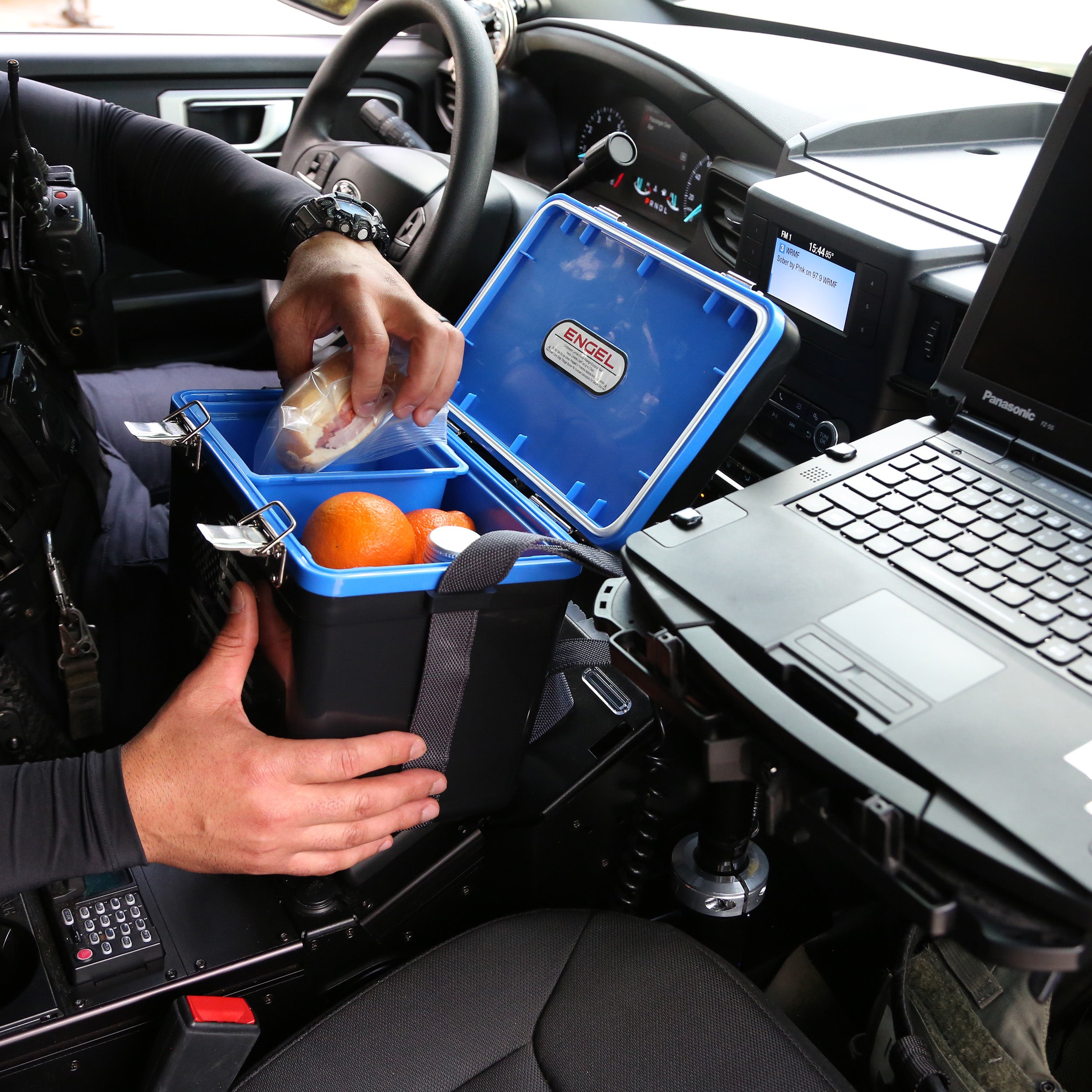 In a vehicle, someone opens an Engel Coolers 7.5Qt Thin Blue Line Lunch Box filled with oranges and food, while a laptop is mounted nearby on a stand, seamlessly blending convenience and functionality.