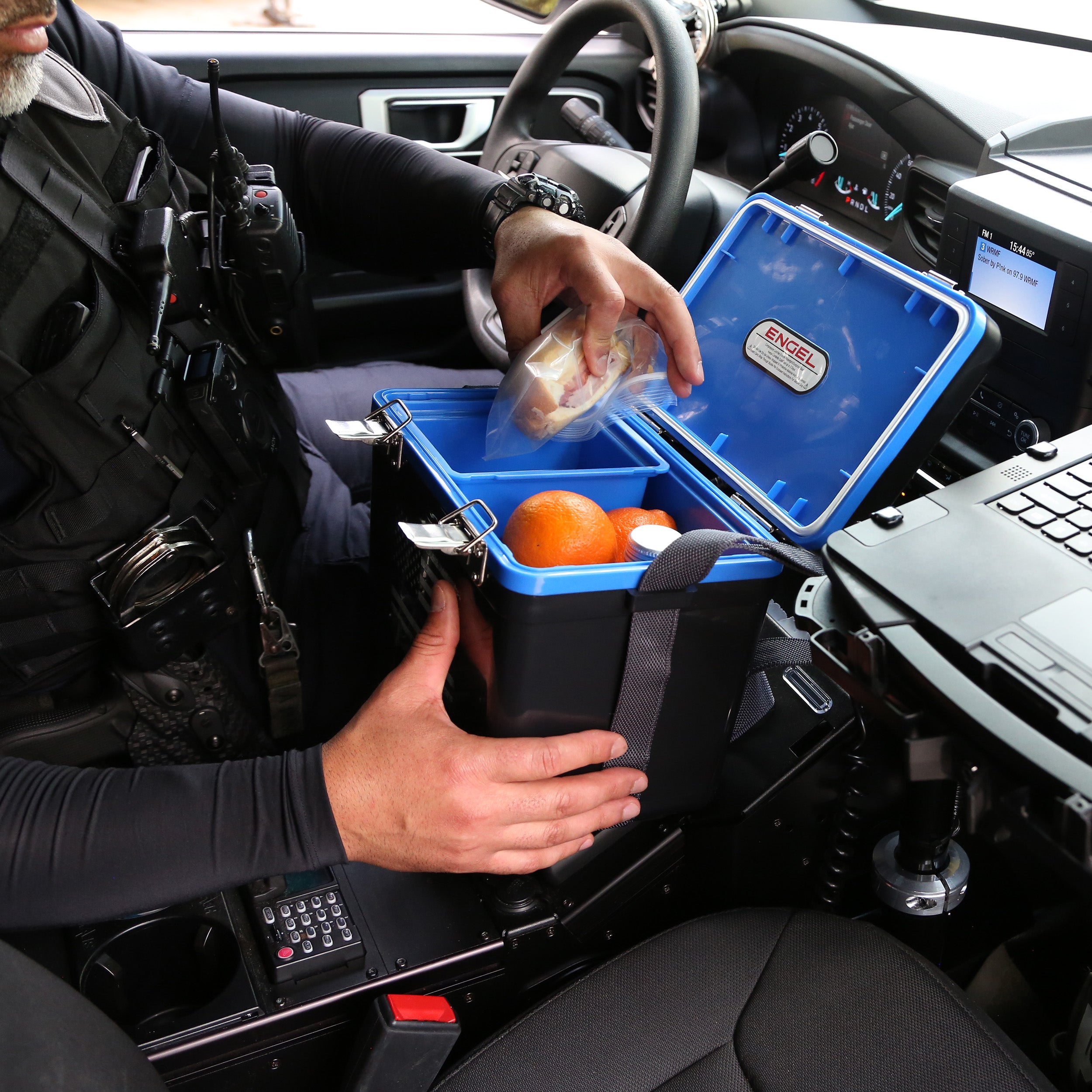 A person in protective gear opens their Engel Coolers 7.5Qt Thin Blue Line Lunch Box in a vehicle, finding fresh oranges and a sandwich inside, while appreciating the design as a tribute to law enforcement.