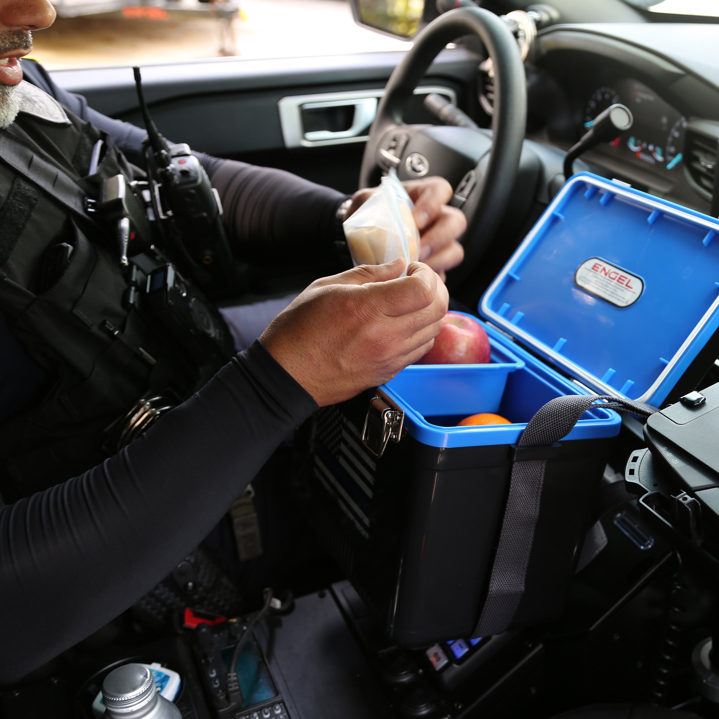 A person in a vehicle, dressed in uniform, places food items into the stylish 7.5Qt Thin Blue Line Lunch Box by Engel Coolers inside the car, embodying dedication similar to a law enforcement tribute.