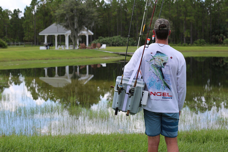A man holding an Engel 30Qt Live Bait Pro Cooler with AP4 XL Rechargeable Aerator, Rod Holders & Stainless Hardware in front of a pond.