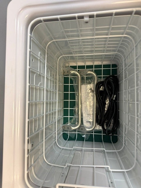Inside the empty white chest freezer, a wire basket holds two plastic-wrapped parts from the Engel Coolers MT45 Platinum Series portable fridge-freezer, along with a coiled black power cord and swing motor compressor components.