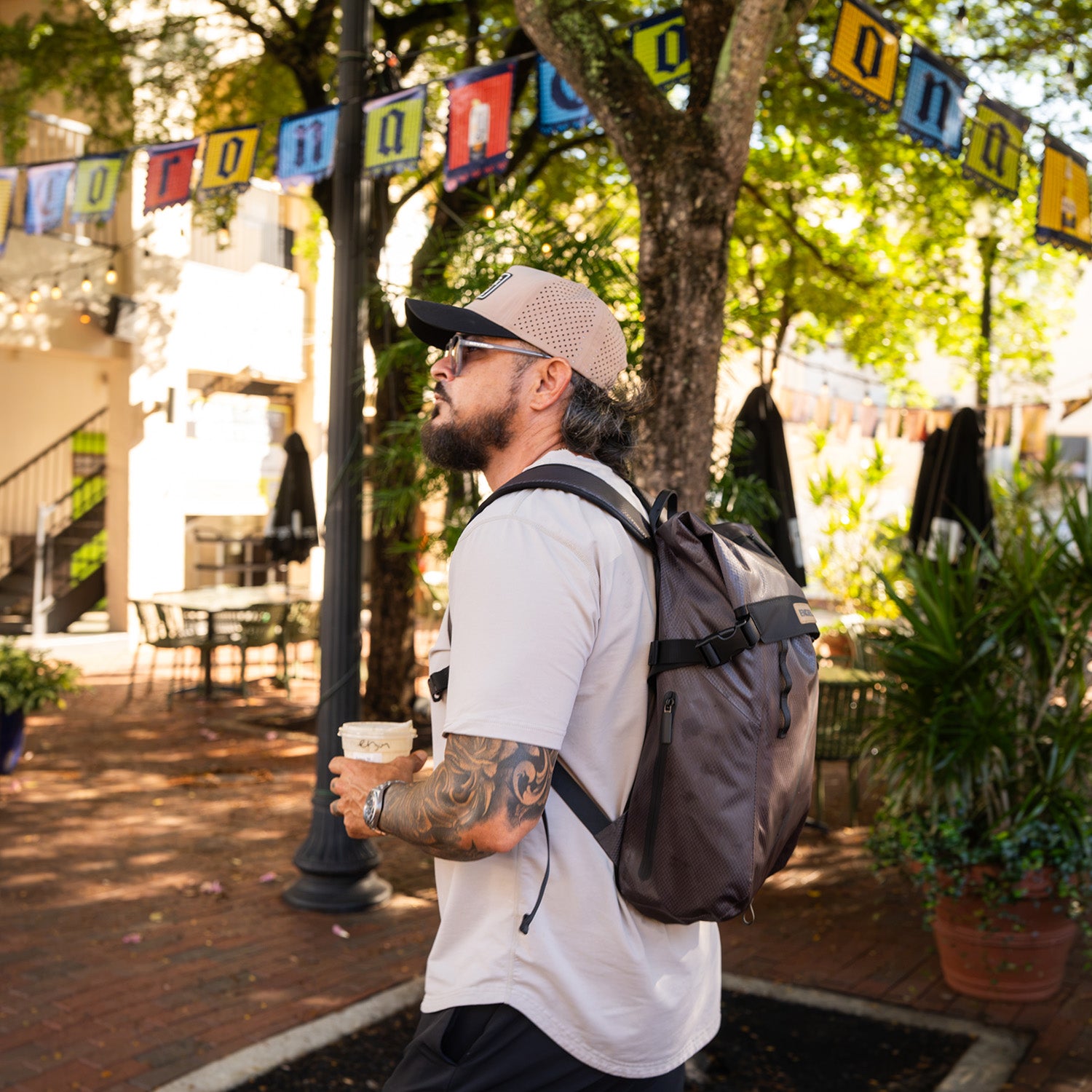 A man wearing a cap and carrying the ENGEL GoBag 20 Qt Backpack Day Bag walks outdoors in a sunny, tree-lined plaza with colorful banners overhead, holding a coffee cup.