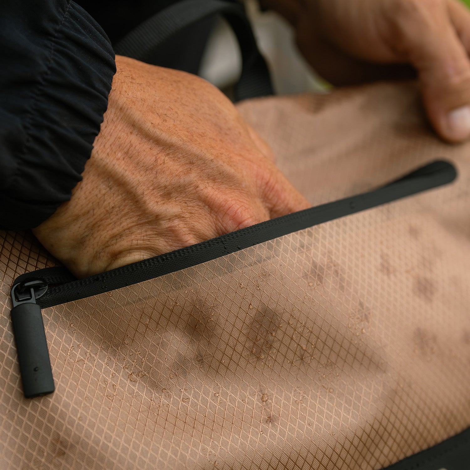 A person opens the zipper of the tan, textured ENGEL Engel GoBag 20 Qt Backpack Day Bag, which has water droplets on its waterproof surface.