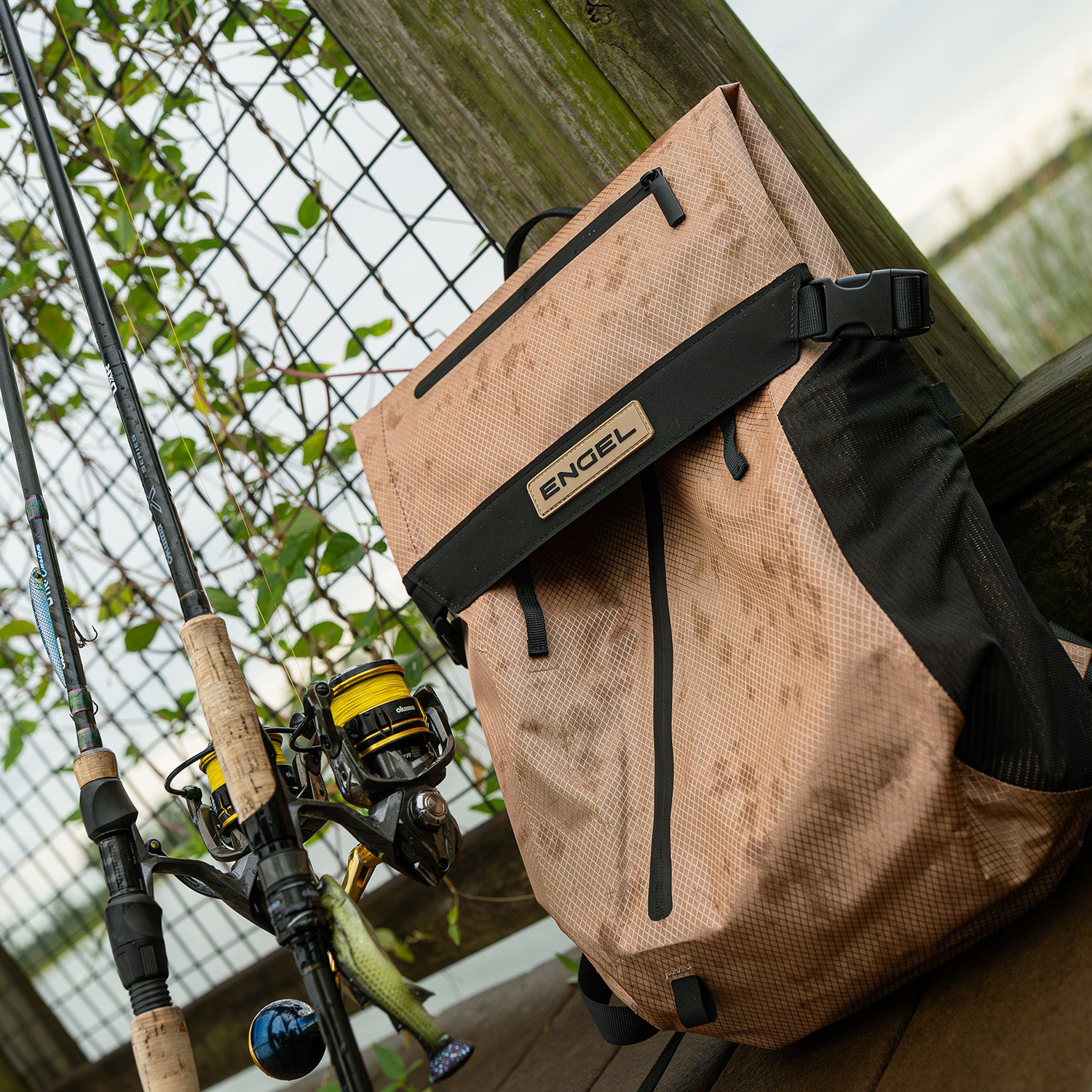 The ENGEL GoBag 20 Qt Backpack Day Bag, featuring black accents, sits next to two fishing rods on a wooden dock, with water and greenery visible in the background.