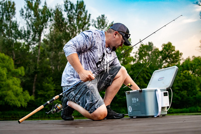 A man kneeling down next to an Engel Coolers 19Qt Live bait Pro Cooler with AP3 Rechargeable Aerator & Stainless Hardware and a fishing rod.