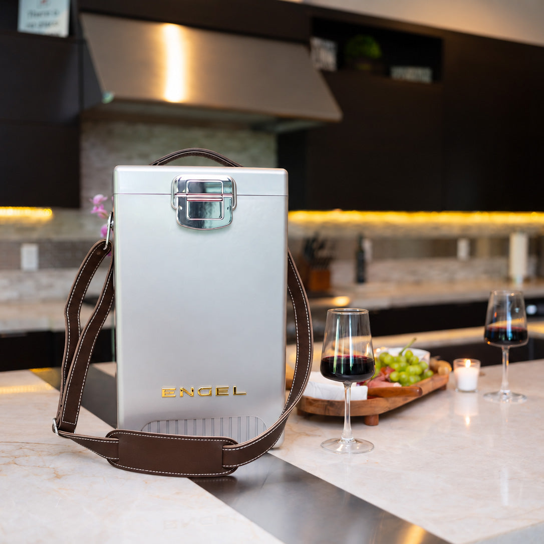 An Engel Reserve Premium Wine Cooler, a tall, silver, rectangular insulated carrier with a brown shoulder strap and a metal latch, stands on a kitchen island with two glasses of red wine and a snack.