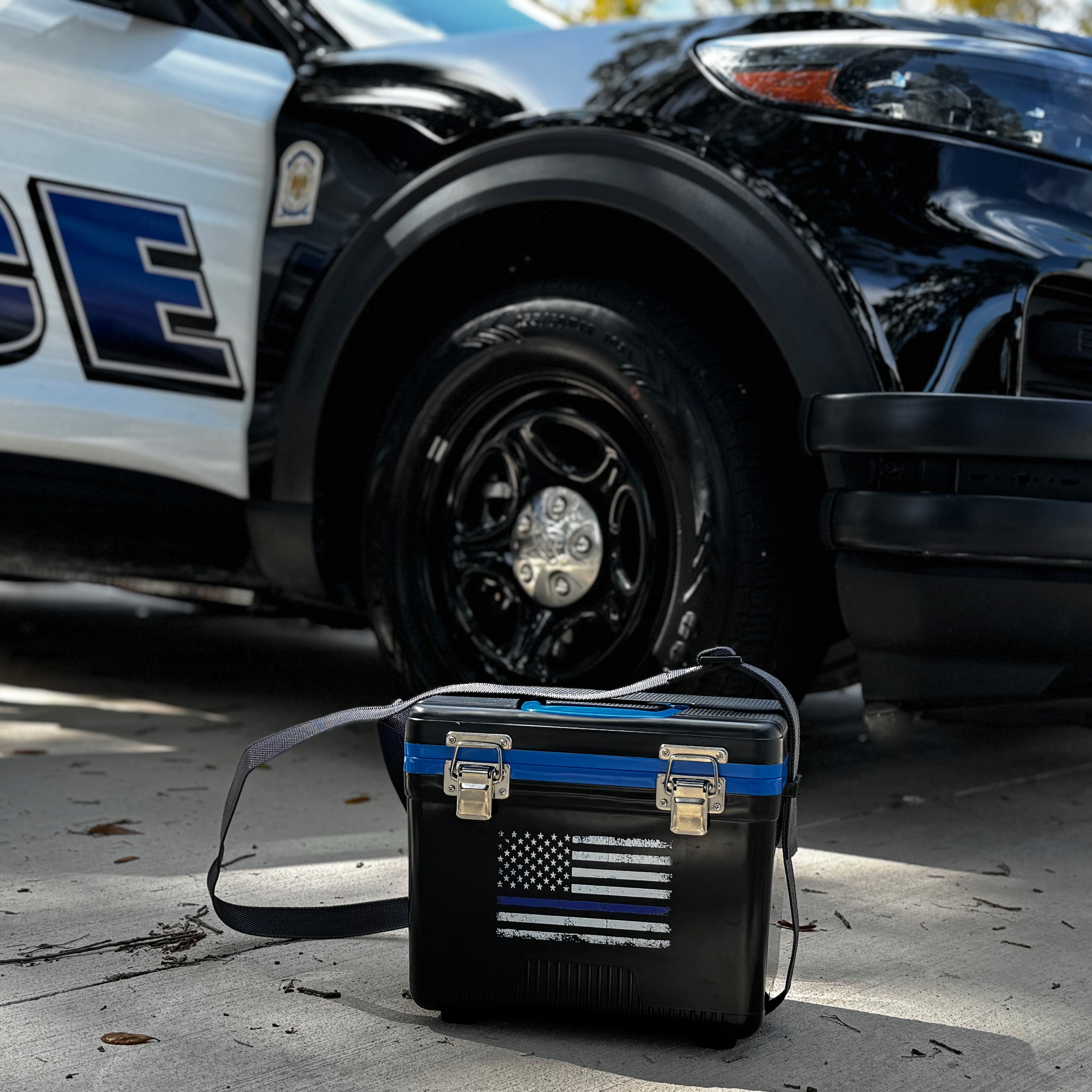 A 7.5Qt Thin Blue Line Lunch Box by Engel Coolers, featuring a Law Enforcement Tribute design, sits on the ground beside the front wheel of a police car.