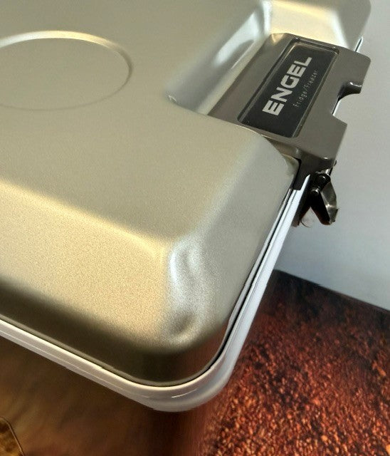 Close-up of a silver Engel Coolers storage container lid, featuring a latch and logo, resting on a brown textured surface—designed to complement the Engel MT45 Platinum Series portable fridge-freezer, renowned for its robust Swing Motor Compressor and dependable performance.