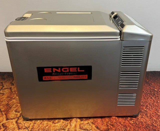 An Engel Coolers MT45 Platinum Series portable fridge-freezer in gray rests on a textured surface, displaying a red and black logo and ventilation slats on the right side. Equipped with Engel’s renowned Swing Motor Compressor, this unit ensures efficient cooling wherever your adventures take you.