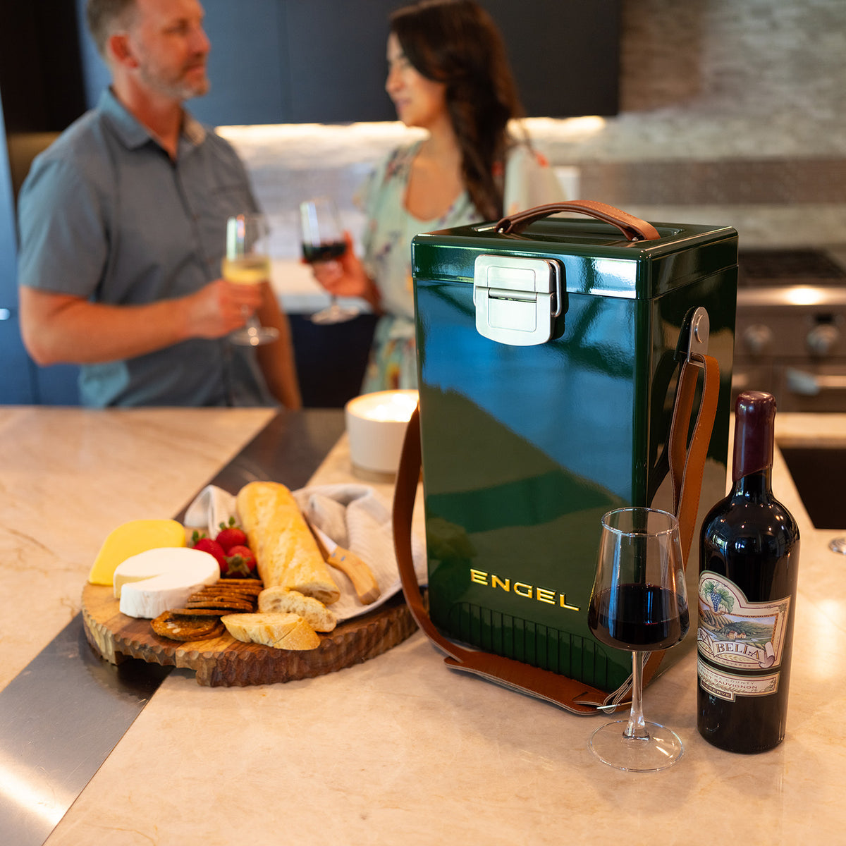 A dark green Engel Reserve Premium Wine Cooler with a leather strap sits on a kitchen counter next to a bottle of wine, a glass, and a wooden board with bread and cheese.