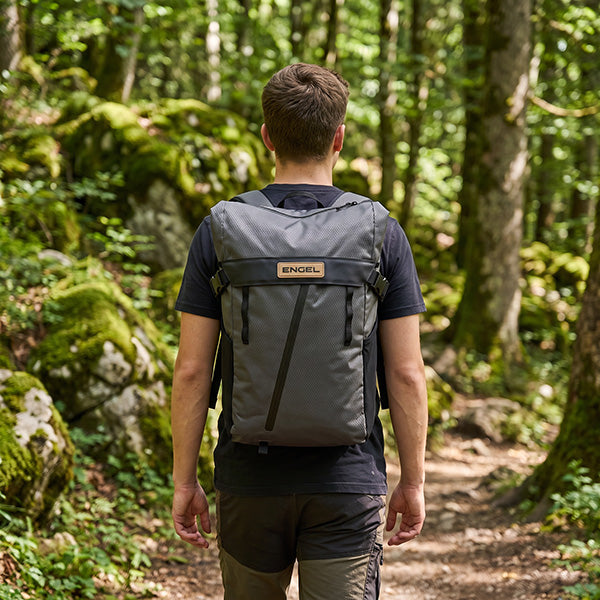 A person with short brown hair stands on a forest trail, seen from behind, wearing a black t-shirt and carrying an ENGEL Engel GoBag 20 Qt Waterproof Backpack in grey, which features a convenient laptop sleeve.