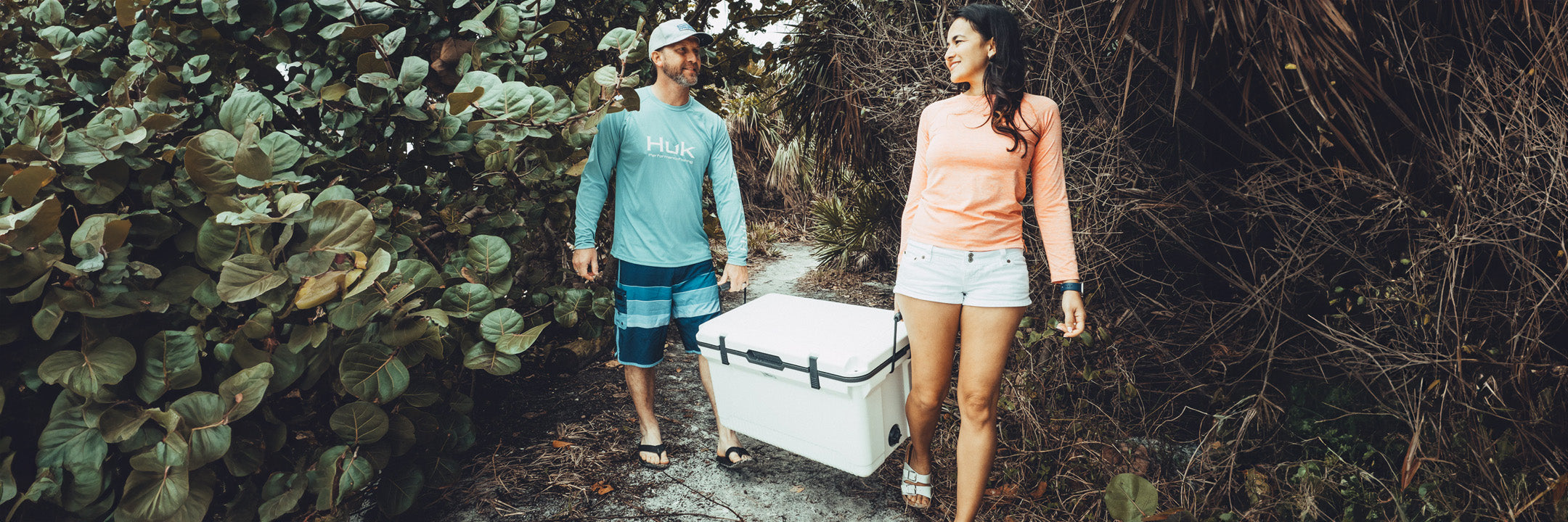 A man and woman carrying a white cooler on a nature trail, smiling at each other