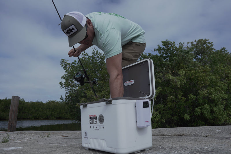 A man leaning over an Engel Cooler equipped with an Engel Extra Large Lithium-ion Rechargeable Live Bait Aerator Pump, holding a fishing rod.