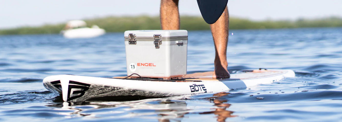 Person standing on a paddle board with a cooler on a calm water surface.