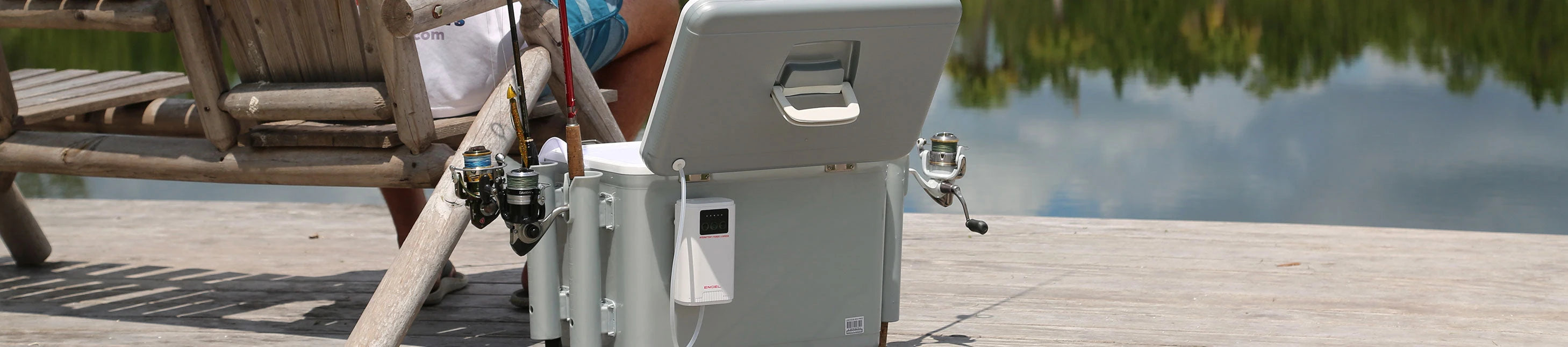 A person sits on a wooden chair on a dock by a lake. There's a fishing cooler with two fishing rods attached.