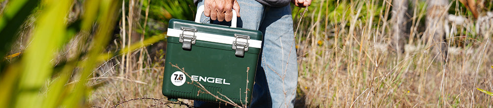 A person carrying a green Engel cooler box with a white handle while walking through tall grass outdoors.