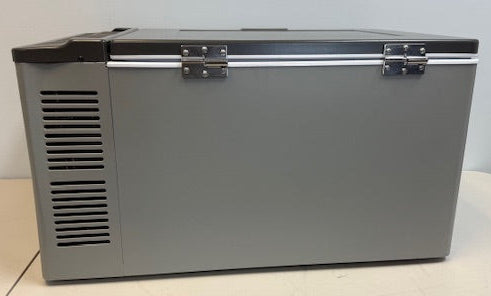 An Engel Coolers MT60 Top Opening Fridge-Freezer (12/24V DC, 110/120V AC) with a gray body, black lid, and metal hinges sits on a beige surface against a light wall, offering precise temperature control for reliable cooling anywhere.