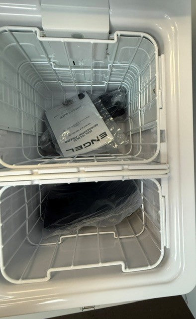 Inside the open Engel Coolers MT45 Combination Platinum Series Top Opening 12/24V DC -110/120V AC Fridge-Freezer, two wire baskets are visible: the upper basket contains a plastic bag and a folded Engel manual, while the lower basket holds a sealed plastic bag. Equipped with an advanced Swing Motor Compressor for reliable cooling, this 45Qt Engel Portable Fridge Freezer is perfect for your outdoor adventures.