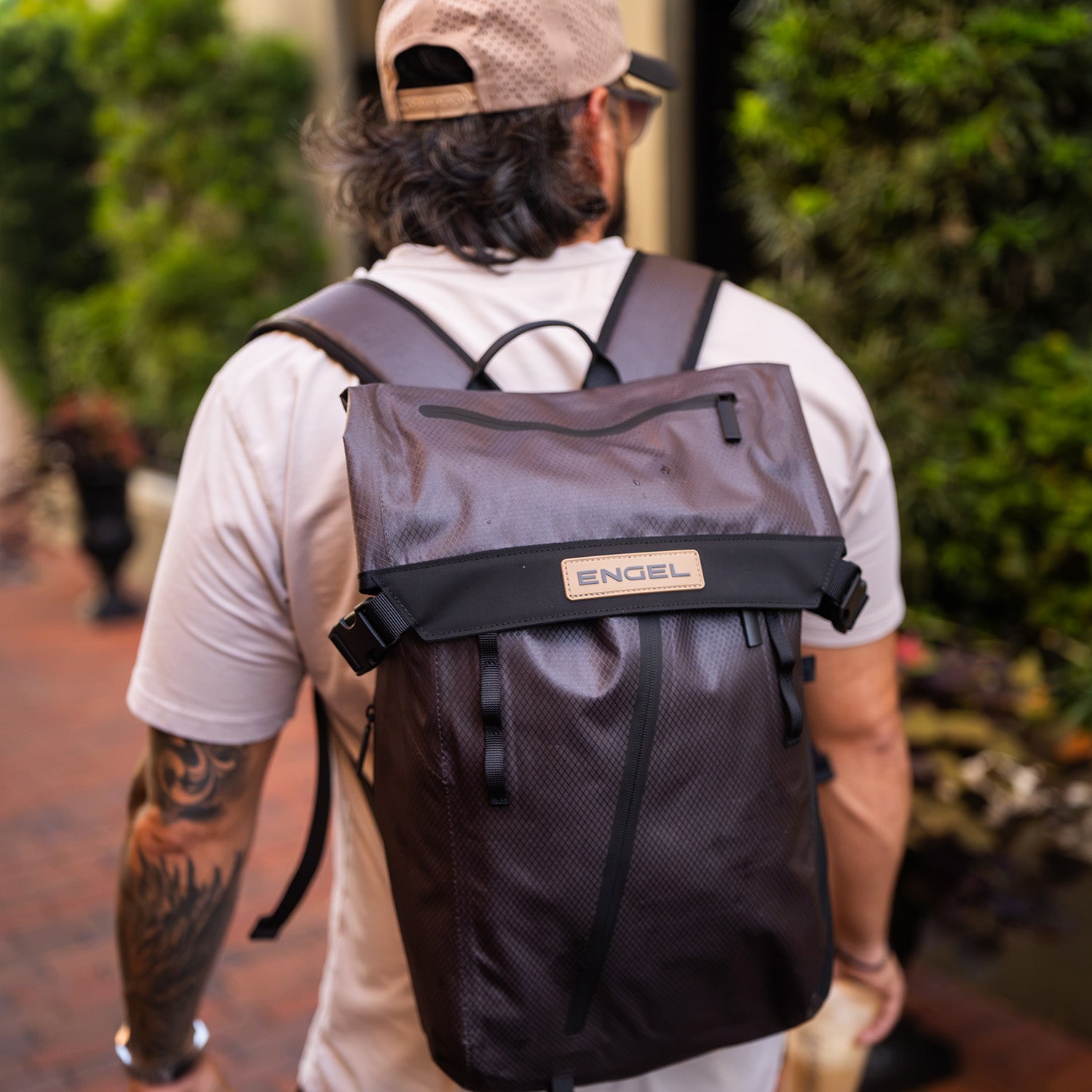 A person with long hair, dressed in a beige cap and shirt, walks outdoors carrying the ENGEL GoBag 20 Qt Backpack Day Bag.
