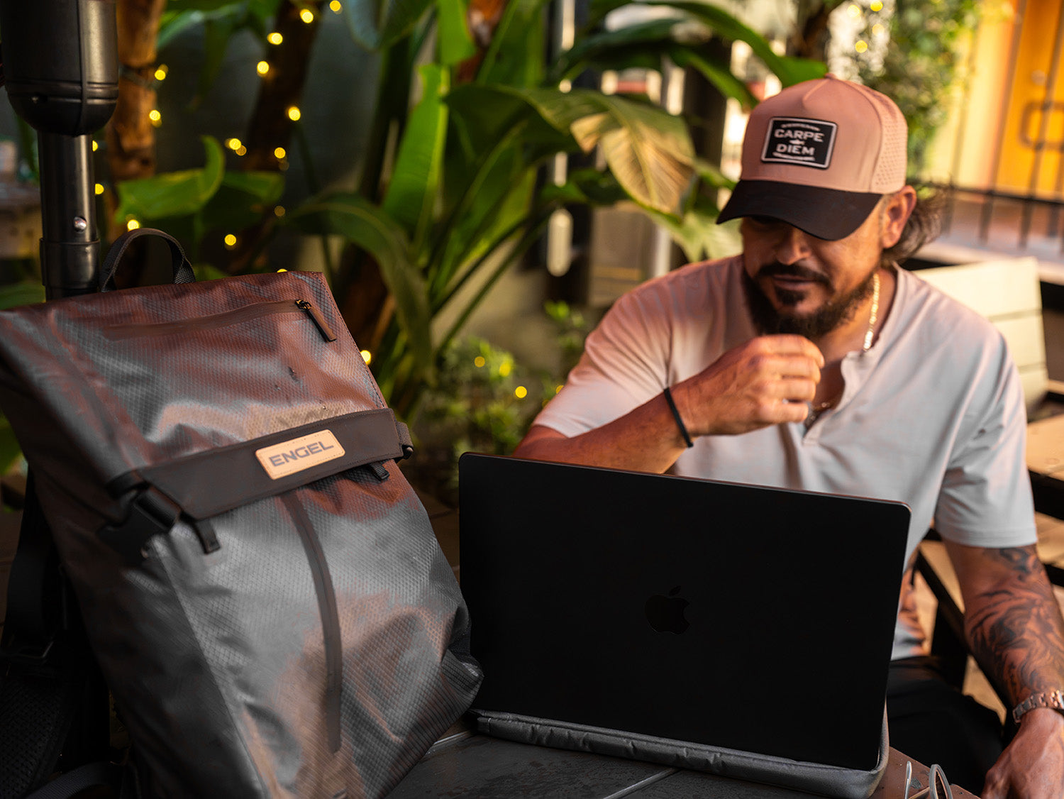 A man in a hat sits at a table outdoors with a laptop and an ultra-light Engel GoBag 20 Qt Backpack Day Bag from ENGEL, surrounded by large green plants and string lights.