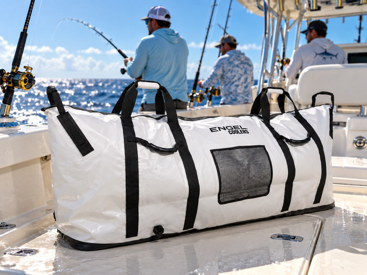 White rectangular Engel Insulated Kill / Fish Bag with a reflective foil liner, black handles, and a mesh pocket on a boat deck, with three people fishing in the background.