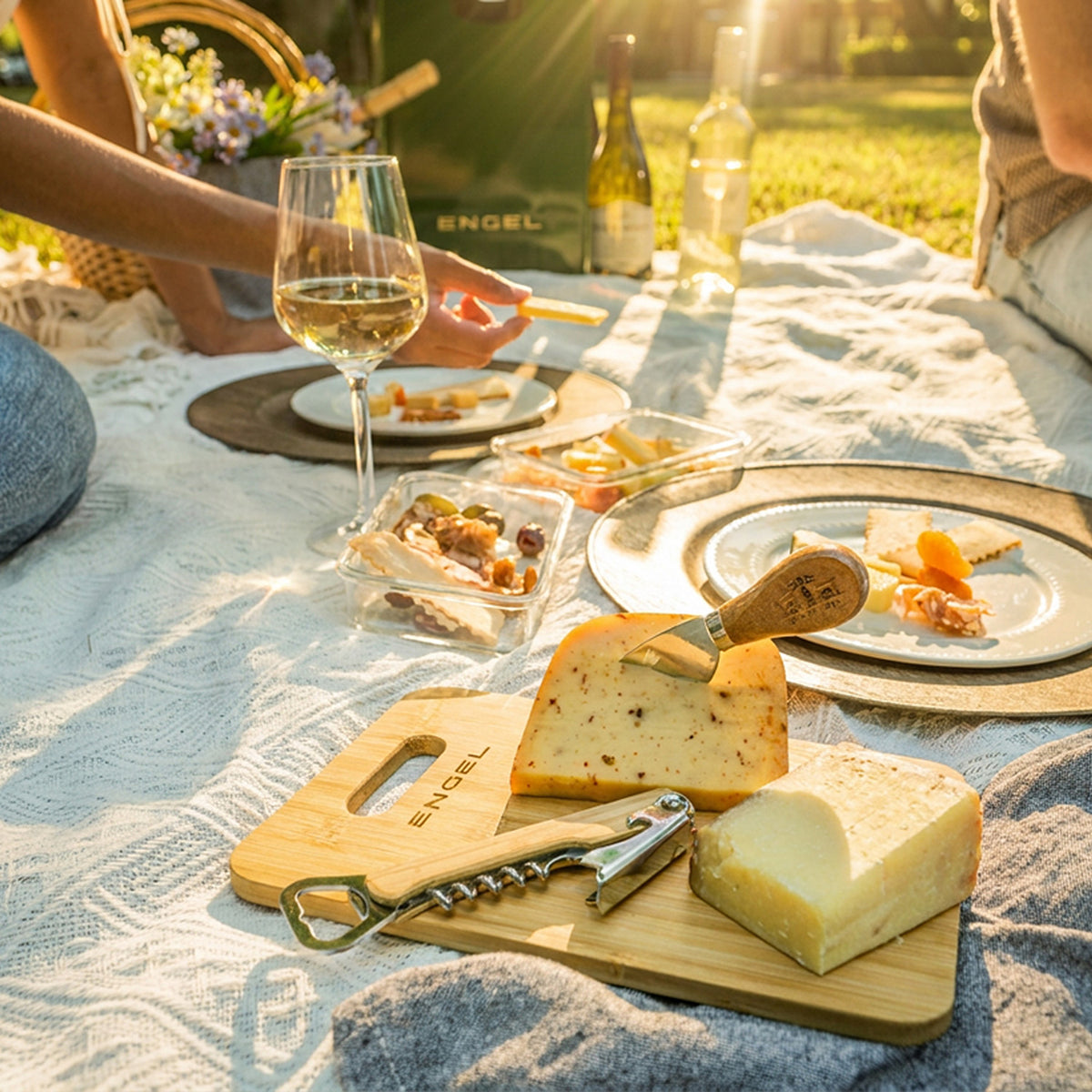 An Engel Reserve Premium Wine Cooler sits on a picnic blanket next to a bamboo cutting board with cheese, a corkscrew, a wine glass, and snacks.