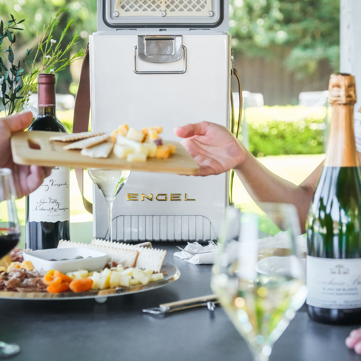 A person hands a wooden board of cheese and crackers across a table featuring an Engel Reserve Premium Wine Cooler, a bottle of wine, and a bottle of champagne.