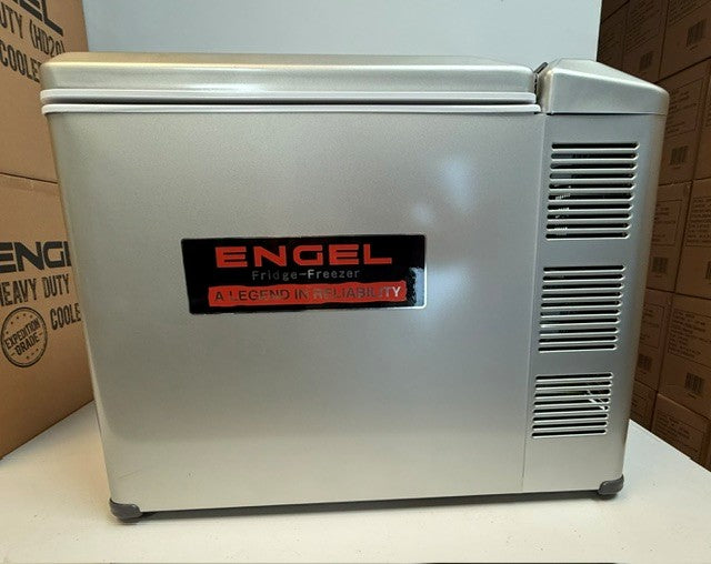 A refurbished Engel MT45 Platinum Series fridge-freezer from Engel Coolers, finished in silver with a black and red logo and side ventilation slots, sits indoors beside cardboard boxes. With its 45-quart capacity, this unit combines reliability and sleek design to meet any indoor storage needs.