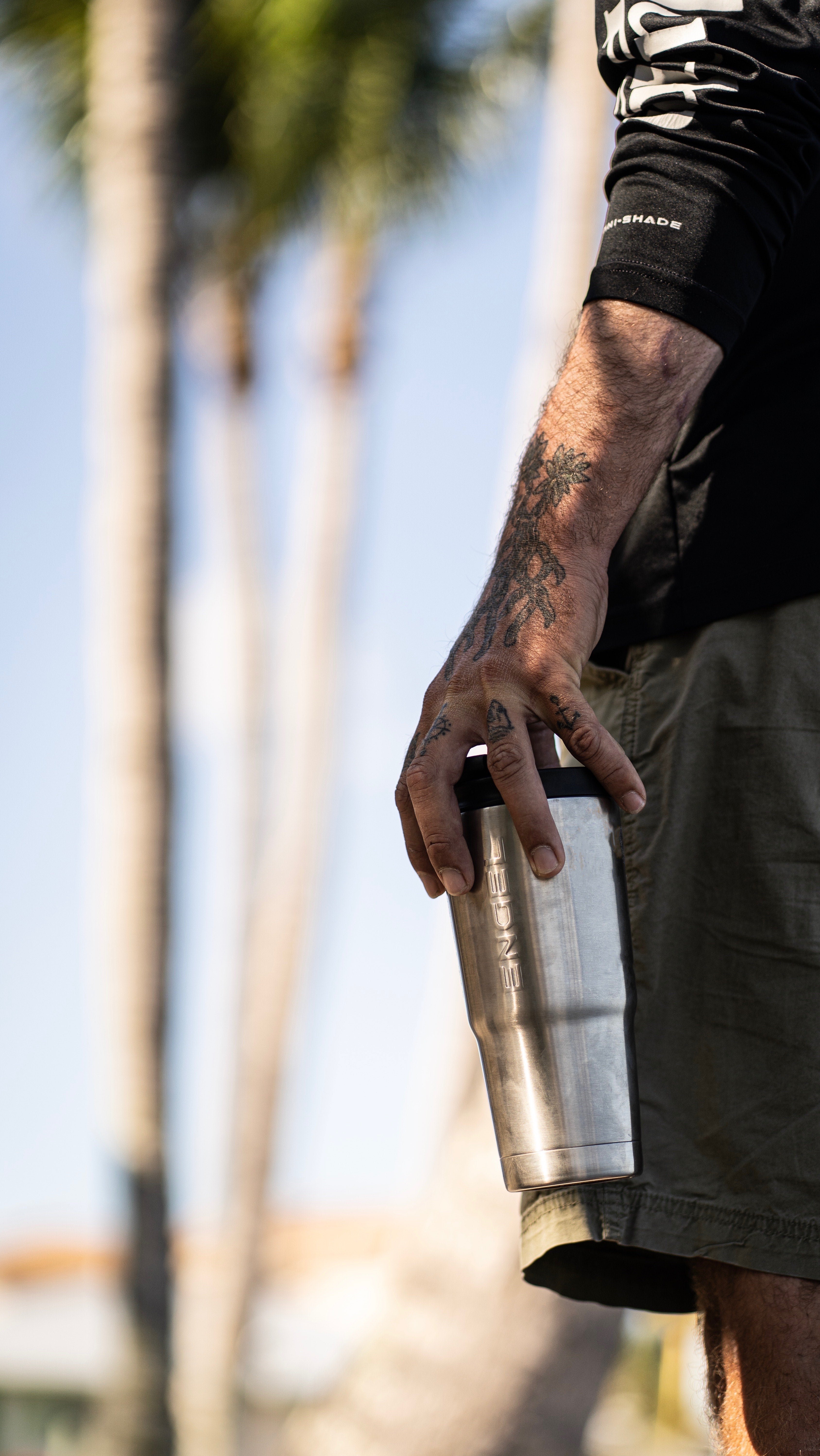 A tattooed individual holds an Engel Coolers 22oz Stainless Steel Vacuum Insulated Tumbler from the Color Collection outdoors, with its shiny surface reflecting the sunlight as palm trees gently sway in the background.