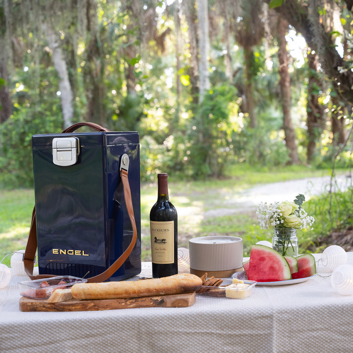 An Engel Reserve Premium Wine Cooler in navy blue beside a bottle of red wine and a picnic spread of bread, fruit, and cheese on a white tablecloth outdoors.