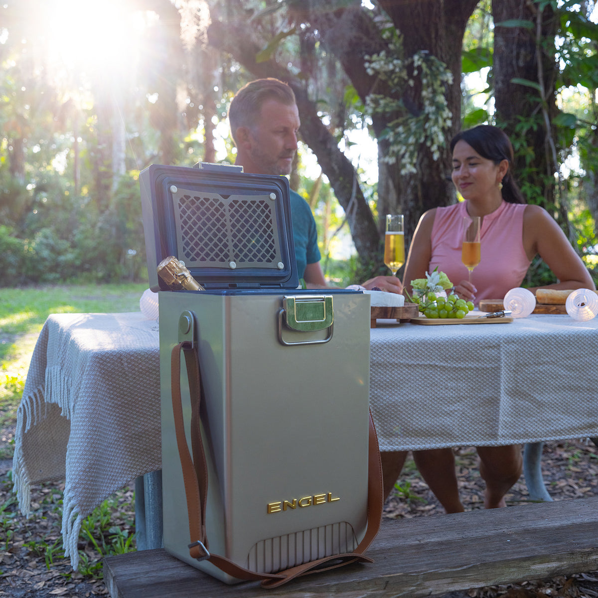A tall, rectangular Engel Reserve Premium Wine Cooler with an open lid containing a bottle sits on a bench in front of a couple dining at a patio table.