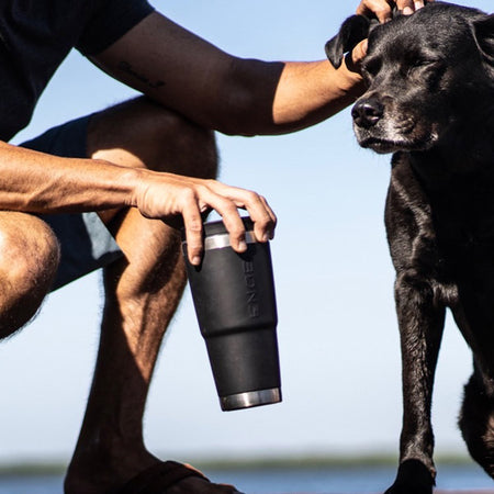 A man petting a black dog while holding a travel mug.