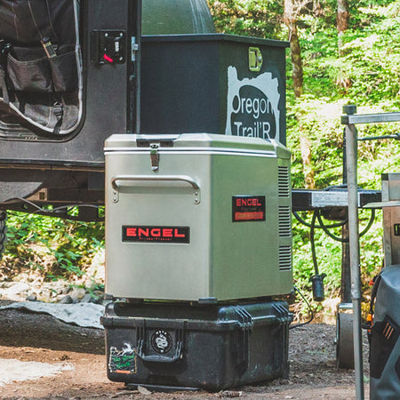 A cooler is parked next to a camper in the woods.