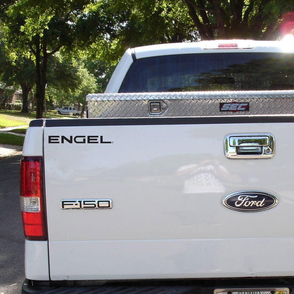 The rear view of a light color vehicle, a white Ford F-150 pickup truck, parked on a residential street, showcases an aluminum tool box in the truck bed and trees in the background. The windows reflect sunlight while a 10" or 15" Engel Coolers Black Vinyl Transfer adds a touch of style to the setup.